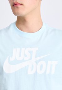 Šviesiai mėlynas medvilninis marškinėlis su dideliu baltu užrašu "TIESIO NEDARYK" ir Nike swoosh logotipu, apvaliu kaklo dizainu ir švelnia tekstūra.