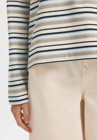 Ριγέ μπλούζα μακρυμάνικη σε μπεζ, καφέ και ανοιχτό γαλάζιο, συνοδευόμενη από παντελόνι σε ανοιχτό μπεζ. Η μπλούζα έχει οριζόντιες ρίγες και άνετη εφαρμογή.