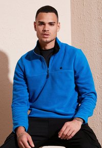 Pullover in pile di colore blu brillante con collo a mezza zip, dettagli neri e un piccolo logo sul petto. Tessuto morbido con maniche lunghe.