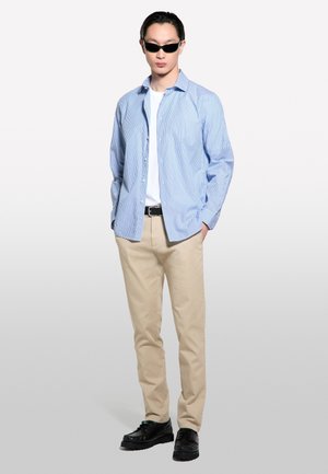 Chemise boutonnée rayée bleu clair portée par-dessus un t-shirt blanc, pantalon kaki et chaussures noires. Les accessoires comprennent des lunettes de soleil noires.
