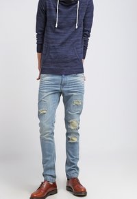 Sweat à capuche marine avec cordons blancs, associé à un jean bleu clair déchiré avec des accents usés, et des chaussures en cuir marron.