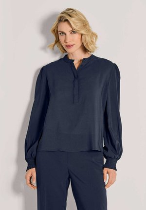 Blonde Frau mit welligem Haar, die eine navyfarbene Bluse mit langen Ärmeln und geknöpften Kragen sowie passende navyfarbene Hose trägt, steht an einer weißen Wand.