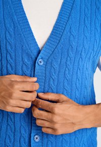 Blå stickad kofta med flätmönster, V-ringning och ribbade kanter. Stängs med en blå knapp framtill.