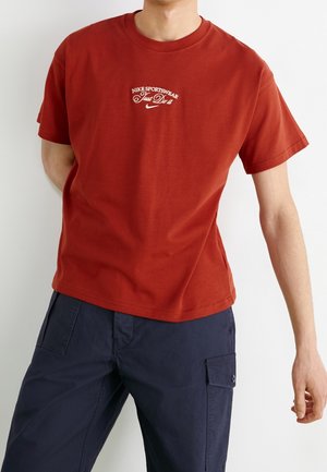 Person trägt ein rotes Nike Sportswear T-Shirt mit dem Text "Just Do It" und dunkelblaue Cargohosen vor weißem Hintergrund.