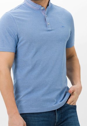 Homme portant un t-shirt henley bleu clair à manches courtes avec une patte de boutonnage à trois boutons et un jean bleu foncé, main dans la poche.