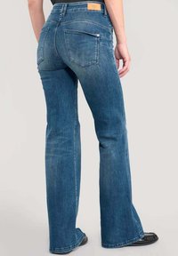 Jeans évasés en denim bleu avec une taille mi-haute, présentant deux poches arrière et une étiquette de marque cousue à la taille.