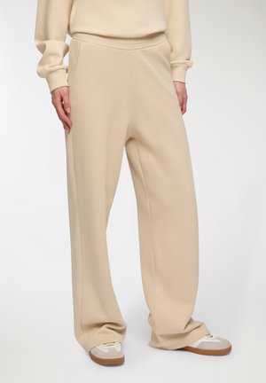 Pantaloni da jogging beige a gamba larga con cintura elastica. Tessuto morbido con una texture liscia. Abbinati a scarpe da ginnastica grigie e bianche.