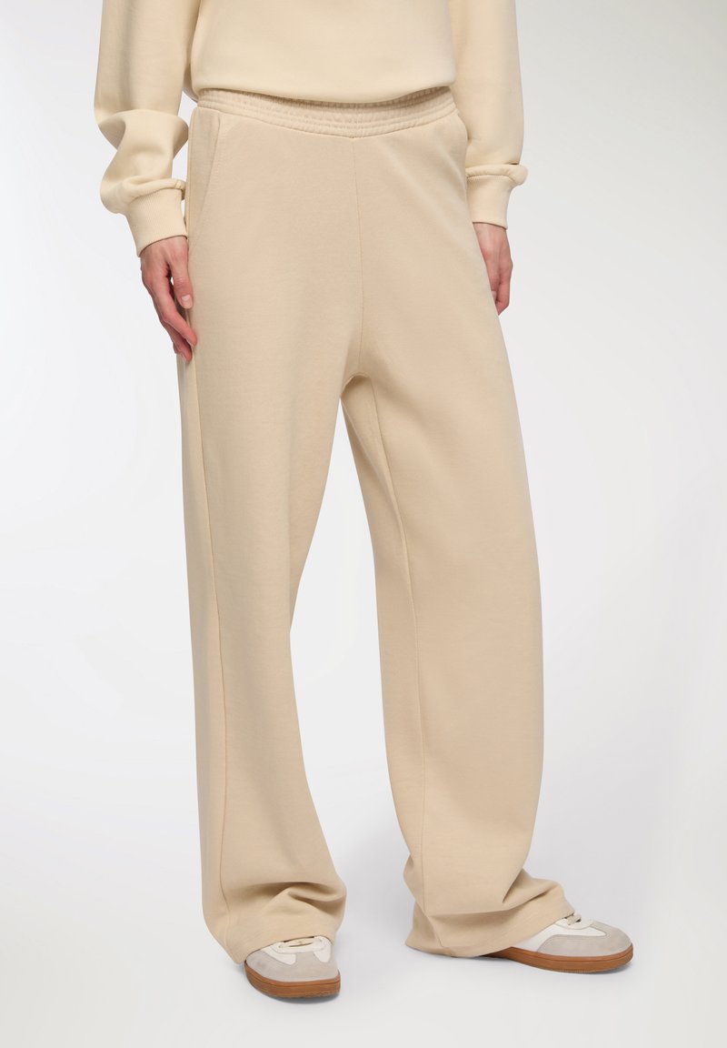 Beżowe, szerokie spodnie dresowe z elastycznym paskiem. Miękka tkanina o gładkiej fakturze. Uzupełnione szarymi i białymi sneakersami.