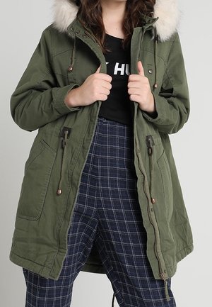 Femme portant un parka vert dézippé avec capuche en fourrure, chemise noire et pantalon à carreaux bleu, tenant les bords avant de la veste avec les deux mains.