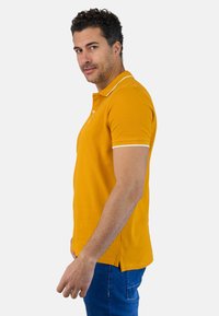 Camisa polo amarilla de manga corta, con acentos blancos en el cuello y las mangas. Hecha de tejido texturizado, muestra un detalle de abertura lateral.