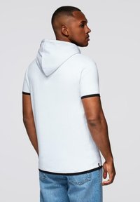 Mladý muž stojící bokem, oblečený v bílém krátkém rukávu s kapucí s černými detaily a modrými džíny, proti jednolitému světlému pozadí.