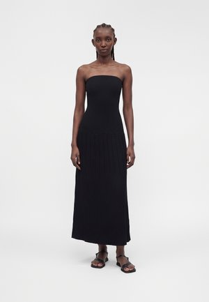Mujer con cabello trenzado que lleva un vestido negro largo sin tirantes plisado y sandalias negras abiertas de punta, de pie frente a un fondo blanco.