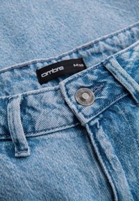 Jeans de mezclilla azul claro con una textura desgastada, presentan un botón de metal y una etiqueta negra con el texto "ombre". Primer plano del área de la cintura.