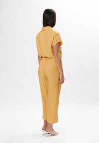 Geel utiliteitsoverall met korte mouwen, elastische taille en cropped pijpen, gecombineerd met witte sandalen met bandjes, gladde stoftextuur.