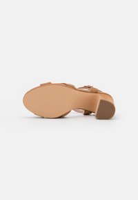 Tan suede heeled sandal with an open toe, ankle strap, and a textured beige rubber sole. Features a slender block heel.