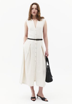 Donna che indossa un abito midi bianco senza maniche con bottoni, cintura nera intrecciata, tiene una borsa a tracolla nera intrecciata e indossa sandali neri con tacco.