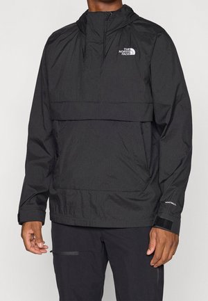 Mężczyzna nosi czarną kurtkę z kapturem North Face WindWall z przednią kieszenią i regulowanymi paskami na nadgarstki.