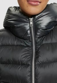 Musta пуховик korkealla kauluksella. Varustettu etuvetoketjulla ja tikatulla kuviolla, valmistettu kiiltävästä, sileästä materiaalista.