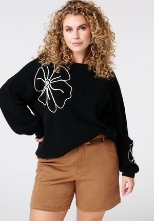 Zwarte gebreide trui met witte bloemenborduursels, gecombineerd met bruine suède shorts. De trui heeft een losse pasvorm en brede mouwen.
