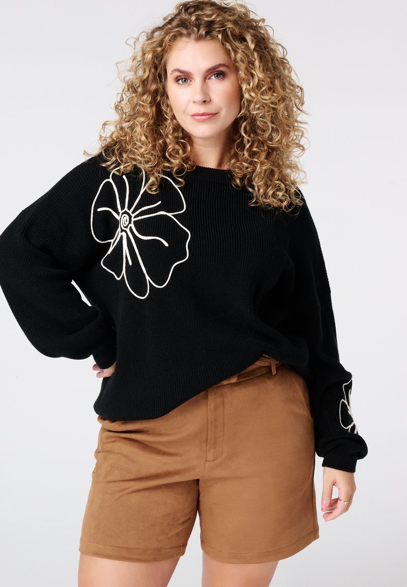 Zwarte gebreide trui met witte bloemenborduursels, gecombineerd met bruine suède shorts. De trui heeft een losse pasvorm en brede mouwen.