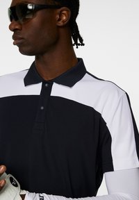 Svart och vit färgblockerad polo skjorta med krage. Har en knappslå och slät tygkvalitet. Vit ärm synlig.