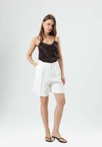 Zwarte satijnen topje met kanten rand, gecombineerd met witte plissé shorts met hoge taille. Model draagt zwarte teenslippers en staat tegen een effen achtergrond.