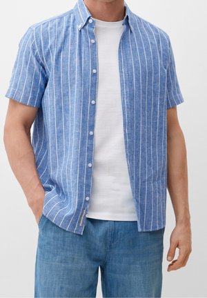 Mann trägt ein blau-weiß gestreiftes Kurzarmhemd mit vertikalen Streifen, offen über einem weißen T-Shirt, dazu blaue Jeanshose, Hände entspannt.