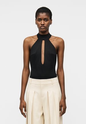 Γυναίκα που φοράει μαύρο αμάνικο τοπ με λαιμόκοψη halter και μπροστινό άνοιγμα σε σχήμα κλειδιού, καθώς και μπεζ παντελόνι με πιέτες, φαρδύ και ψηλόμεσο, σε λευκό φόντο.