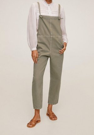 Kvinde iført olivengrøn denim-overalls, hvid bluse med lange ærmer og brune flade sandaler, stående mod neutral baggrund.