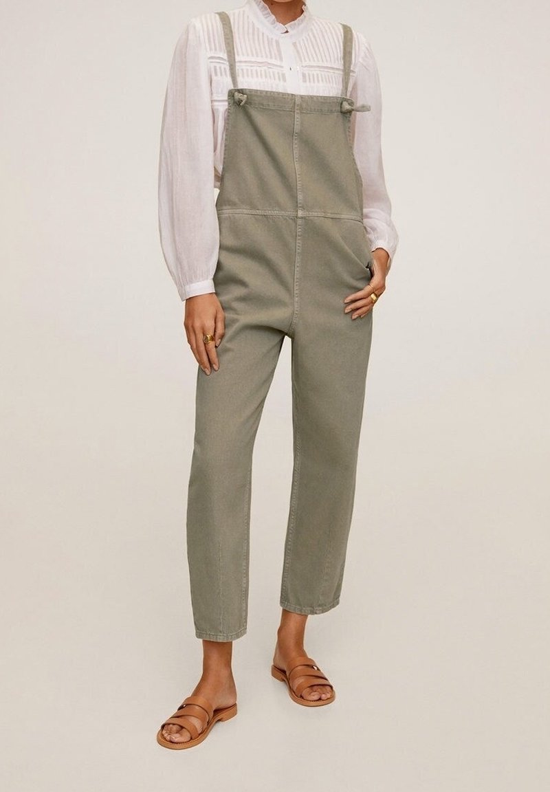 Femme portant une salopette en denim vert olive, un chemisier blanc à manches longues et des sandales plates marron, debout devant un fond neutre.