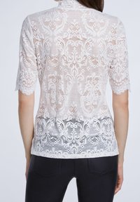 Haut en dentelle écru avec motif floral, manches courtes, col montant et ourlet festonné, superposé sur un bas uni.