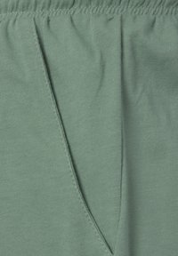 Zaļš kokvilnas audums ar mīkstu tekstūru, kas aprīkots ar elastīgu vidukli un divām sānu vīlēm, izceļot tā ikdienišķo dizainu.