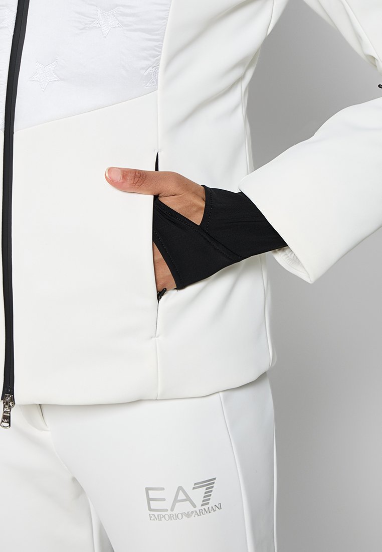 Giacca bianca con zip, con motivo trapuntato a stelle sulle spalle, maniche nere e tasche frontali; include il logo EA7 sulla parte inferiore anteriore.