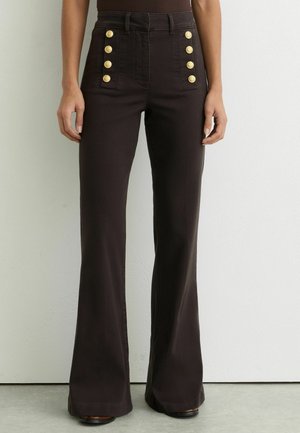 Pantaloni a zampa di elefante marrone scuro a vita alta con due file verticali di quattro bottoni dorati ciascuna sulle tasche anteriori, indossati con una maglietta infilata dentro.