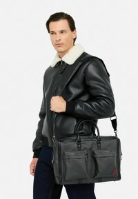 Schwarze Lederhandtasche mit zwei Fronttaschen und rotem Logo, ausgestattet mit zwei Griffen und einem abnehmbaren Schultergurt. Glatte Textur, strukturierte Form.