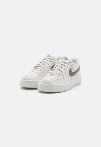 Pár bílých tenisek Nike Air Force 1 se šedými logy swoosh, bílými tkaničkami a perforovanými špičkami na jednobarevném pozadí.