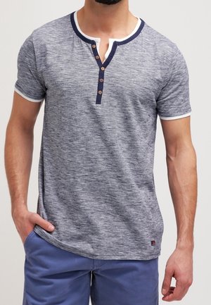 Mies, jolla on harmaa lyhythihainen henley-paita laivastonsinisillä somisteilla ja napeilla, yhdistettynä sinisiin shortseihin, seisoo käsi taskussa.