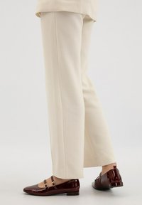 Beżowe spodnie eleganckie z teksturowanym wykończeniem, w połączeniu z ciemnoczerwonymi lakierowanymi butami Mary Jane z paskami na kostkę i niskim obcasem.