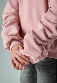 Růžová mikina s objemovými, nařasenými rukávy a žebrovanými manžetami, v kombinaci se šedými džíny. Hladká textura a uvolněný střih.