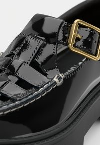 Sapato de couro envernizado preto com design trançado, detalhe de fivela dourada, sola preta robusta e costura branca ao longo da borda.