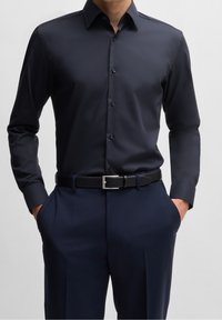Camicia blu navy con bottoni, dalla texture liscia, maniche lunghe e colletto strutturato, abbinata a pantaloni scuri e cintura nera con fibbia argento.