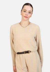 Beige lange mouw top met een ronde halslijn, gecombineerd met crèmekleurige broeken, in de taille aangesnoerd met een bruine riem met een gouden gesp.