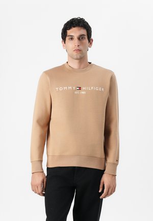 Bruine sweatshirt met ronde hals, voorzien van het "TOMMY HILFIGER" logo en de tekst "EST. 1985" in het wit. Geribbelde manchetten en tailleband. Donkere jeans.