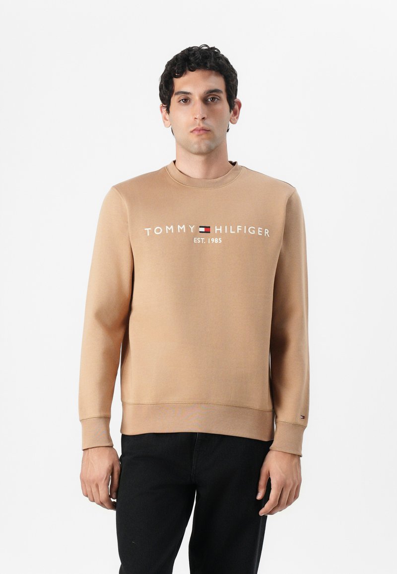 Brun sweatshirt med rund hals, der har "TOMMY HILFIGER" logo og teksten "EST. 1985" i hvid. Ribbet manchetter og talje. Mørke jeans.