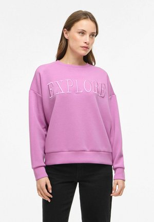 Jonge vrouw die een lavendelkleurige trui met de tekst "EXPLORE" en een zwarte broek draagt, staand tegen een effen witte achtergrond.