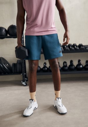 Mężczyzna w różowej koszuli i niebieskich szortach trzyma hantlę, stojąc na siłowni z kettlebellami i ciężarami na regałach za nim.