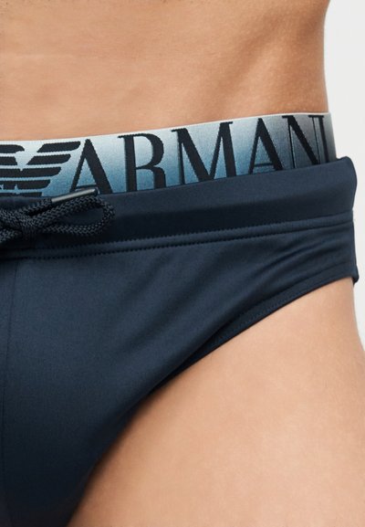 Η μέση ενός άνδρα που φοράει μπλε σκούρο μαγιό με μαύρο κορδόνι, αποκαλύπτοντας μέρος της γκρι μέσης εσωρούχου Armani με λογότυπο και έμβλημα αετού.