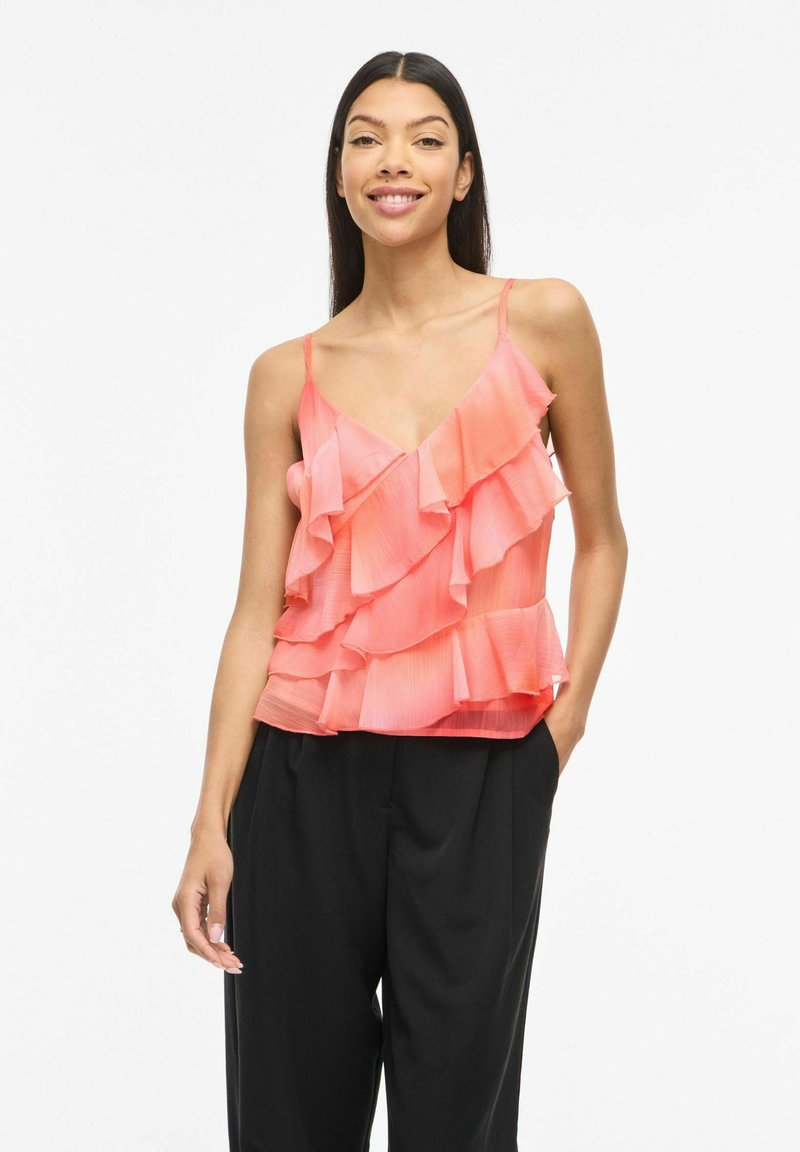 Femme souriante aux longs cheveux foncés portant un haut rose sans manches à volants et un pantalon noir ample, debout devant un fond blanc.