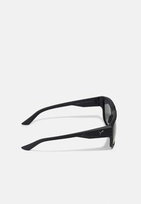 Ochelari de soare negri cu un design aerodinamic, fabricați din plastic. Prezintă lentile plate, brațe groase și un logo discret lângă balama.