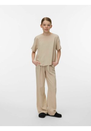 Młoda osoba stojąca z rękami w kieszeniach, ubrana w beżową, luźną koszulę i spodnie oraz czarne buty, na tle jednolitego białego tła.